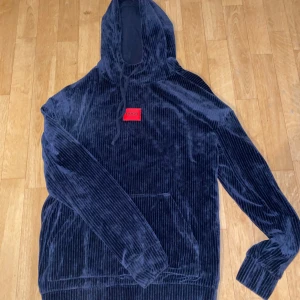 Blå manchester hoodie från Hugo - Snygg blå manchester hoodie från Hugo med en röd logga på bröstet. Tröjan har en stor ficka framtill och en justerbar huva med dragsko. Perfekt för en avslappnad stil. Helt oanvänd. Skriv för minsta fundering.