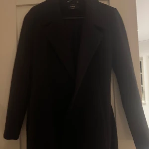Svart kappa från ONLY - Elegant svart kappa från ONLY i storlek XS. Den har en klassisk design med knytbälte i midjan och långa ärmar. 