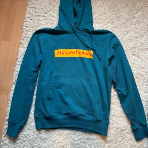 Helmut Lang hoodie - Säljer denna Helmut Lang hoodien i turkos färg. Hoodien är tjock och varm och har ett gult tryck på magen med Helmut Lang på. Nypris för en Helmut Lang hoodie ligger på ca 3000sek. 