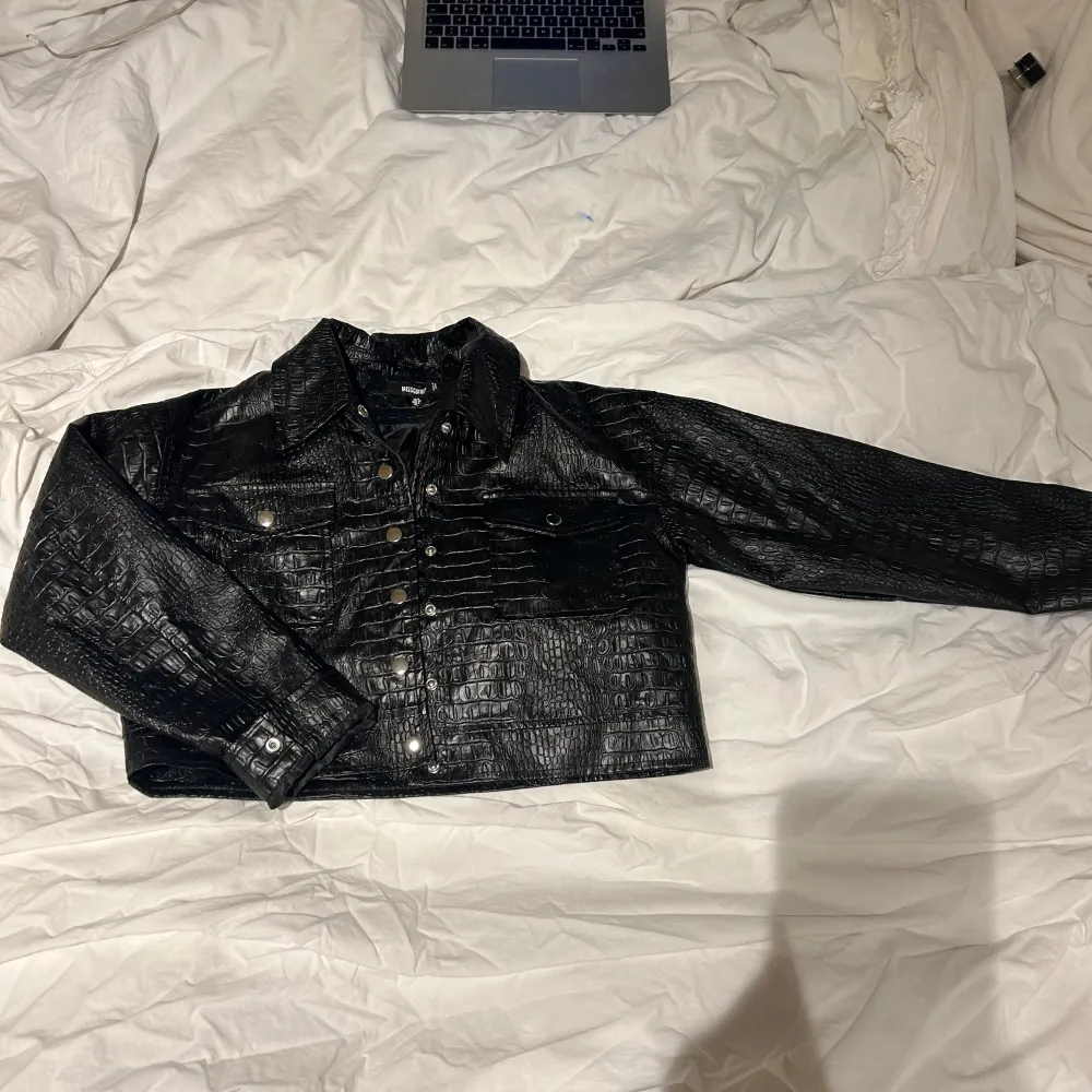 Snygg svart croppad jacka från Missguided med krokodilpräglat mönster och knappar framtill. Jackan har långa ärmar och två bröstfickor med knappdetaljer. Perfekt för en trendig look!. Takit.