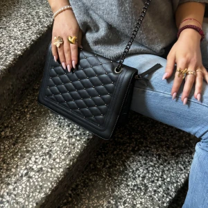Svart quiltad axelväska - Snygg svart axelväska med quiltad design och kedjerem som går att ha längre eller kortare om man vill det . Perfekt för att ge en stilren touch till din outfit. Väskan har en elegant och tidlös look.
