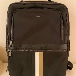 Svart ryggsäck från Boss - Snygg svart ryggsäck från Boss med stilrena detaljer. Väskan har flera dragkedjor och justerbara axelremmar för bekväm passform. Perfekt för både vardag och arbete.