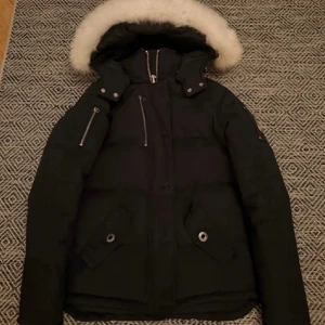 Svart dunjacka från Moose Knuckles - Snygg svart dunjacka från Moose Knuckles med avtagbar huva och vit pälsdetalj. Jackan har flera praktiska fickor med dragkedja och knappar. Perfekt för kalla vinterdagar.