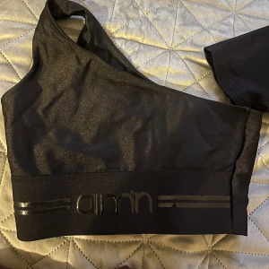 Svart oneshoulder topp från Aim'n - Snygg svart oneshoulder topp från Aim'n med glittrig finish. Perfekt för en trendig look. Den har en bred resår med logotypdetalj nedtill.