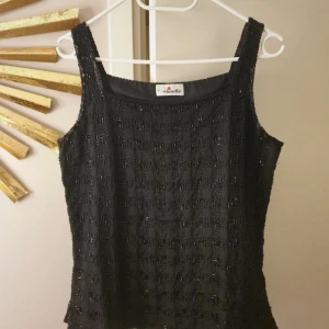 Svart pärlbroderad topp från Elinette - Elegant svart topp från Elinette med vackra pärlbroderier. Denna ärmlösa topp har en fyrkantig halsringning och är perfekt för en stilfull look.