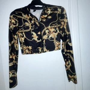 Svart och guldmönstrad croppad tröja - Snygg croppad tröja i svart med ett elegant mönster i guld. Llånga ärmar och en dragkedja framtill. Perfekt för att ge en stilfull touch till din outfit.