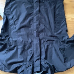 BUDA: Svart regn-/ springjacka från Didriksons utan luva - Säljer en svart regnjacka från Didriksons, perfekt för regniga dagar. Jackan är 100% vattentät och tillverkad i polyester. Den har knappar och en stilren design.