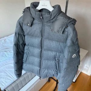 Grå dunjacka från Moncler - Säljer en snygg grå dunjacka från Moncler med dragkedja och hög krage. Jackan har långa ärmar och en broderad Moncler-logga på ärmen. Perfekt för kyliga dagar med sin quiltade design.