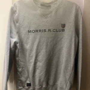 Grå sweatshirt från Morris R. Club - Säljer en stilren grå sweatshirt från Morris R. Club med tryck på bröstet och ett litet emblem med brittisk flagga. Tröjan har långa ärmar och rund halsringning. Perfekt för en avslappnad stil.