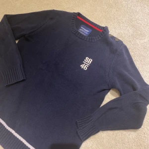 Mörkblå tröja från Sebago - Säljer en snygg mörkblå tröja från Sebago med broderad text 'SB 46' på bröstet. Tröjan har långa ärmar och knappar vid axeln för en stilren look. Perfekt för en avslappnad och stilfull vardag.