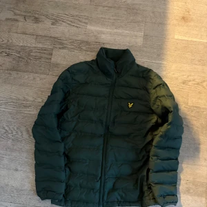 Grön pufferjacka från Lyle & Scott - Säljer en snygg grön pufferjacka från Lyle & Scott med dragkedja och deras ikoniska logga på bröstet. Perfekt för kyliga dagar med sin quiltade design och långa ärmar.