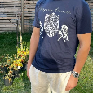 Mörkblå t-shirt från Dalla Dolce - Snygg mörkblå t-shirt från Dalla Dolce med ett elegant tryck i vitt. Texten 'Élégance Éternelle' och ett stiliserat emblem med hästar ger en exklusiv känsla. Perfekt för en avslappnad men stilren look.