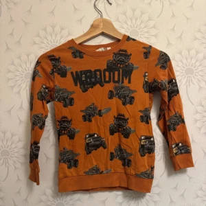 Orange långärmad tröja med biltryck - Cool orange tröja med lång ärm och tryck av svarta bilar och texten 'VROOM' på framsidan. Perfekt för bilälskaren!