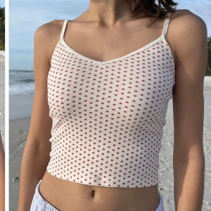 Vitt linne med röda hjärtan - Söt vit linne med små röda hjärtan över hela tyget. Den är ärmlös med smala axelband och har en croppad passform. Det är one size och linnet är från Brandy Melville 