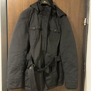 Svart parkajacka med bälte - Snygg svart parkajacka från Esprit med justerbart bälte i midjan och praktisk huva. Jackan har långa ärmar och knappar framtill samt fickor med lock. Perfekt för kyligare dagar.