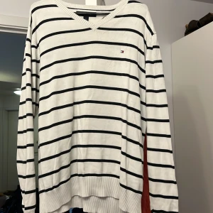 Vit och svart randig tröja från Tommy Hilfiger - Säljer en stilren vit tröja med svarta ränder från Tommy Hilfiger. Tröjan har en v-ringad hals och långa ärmar, perfekt för en avslappnad look. Den ikoniska loggan är broderad på bröstet.