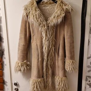 Snygg pennylane kappa med pälsdetaljer - Säljer en snygg beige varm kappa med fluffiga pälsdetaljer på krage, ärmslut och nederkant. Perfekt för kyliga dagar med sin långa design och mysiga känsla. Kappan har en figursydd passform och är helfodrad för extra värme.