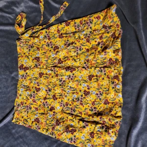 Gul blommig kjol från Bershka - Snygg gul kjol från Bershka med ett färgglatt blommönster. Kjolen har en omlott-design och knytband i midjan för en justerbar passform. Perfekt för att liva upp garderoben med lite färg och mönster.