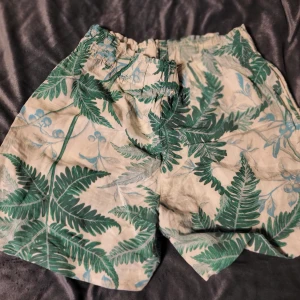 Gröna blommiga shorts i linneblandning - Säljer ett par gröna shorts med blommigt mönster i linneblandning. Perfekta för sommardagar med sin lätta och luftiga känsla. Elastisk midja för extra komfort. Oanvända