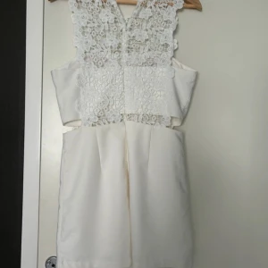 Vit spetsklänning - Elegant vit klänning med vacker spetsdetalj på överdelen. Klänningen är ärmlös och har en kort längd, perfekt för sommarens festligheter. Den har en stilren design med en liten slits framtill.
