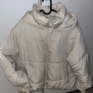 Beige pufferjacka med huva - Säljer en vit pufferjacka med huva. Jackan har en mysig och varm design, perfekt för kyligare dagar. Den har ett par små defekter: Bild 3- en liten reva (hjälper gärna att sy ihop). Bild 4- bläckpenna sträck som inte syns när luvan är nere. Bild 5- mindre fläckar