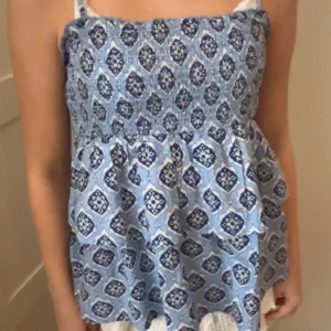 Blå mönstrad top - Söt blå linnetopp med ett detaljerat mönster i vitt och mörkblått. Toppen har smala axelband och en smockad överdel som ger en bekväm passform. Perfekt för varma sommardagar!