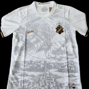 Vit fotbollströja från Nike - Snygg vit fotbollströja från Nike med ett detaljerat grått mönster av en stadssilhuett. Tröjan har korta ärmar och klubbemblem på bröstet. Perfekt för fotbollsfans som vill ha något unikt och stilrent.