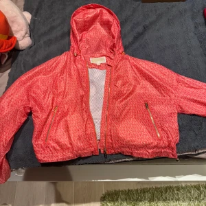 Röd vindjacka från Michael Kors - Snygg röd vindjacka från Michael Kors med ett diskret mönster och dragkedja. Jackan har huva och två praktiska fickor med dragkedjor. Perfekt för blåsiga dagar. Nypris 3000kr