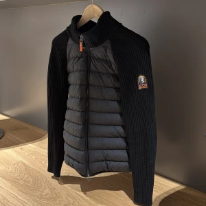 Svart quiltad jacka med stickade ärmar - Snygg svart jacka från Parajumpers med quiltad front och stickade ärmar. Jackan har en dragkedja framtill och en broderad logga på ärmen. Perfekt för kyliga dagar med sin stilrena design och bekväma passform. Priset kan diskuteras!