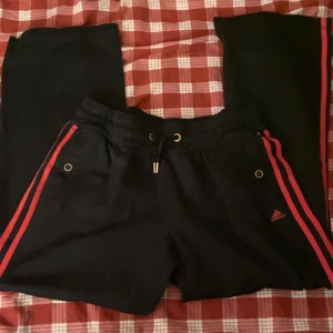 Mjukisbyxor - Snygga svarta träningsbyxor/mjukisbyxor från Adidas med röda ränder längs sidorna. Byxorna har en snörning och praktiska fickor. Perfekta för en sportig look. Står ingen storlek men passar S och M.