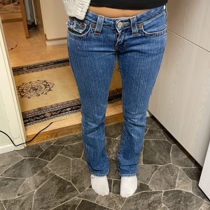 Blå jeans från True Religion - Snygga blå jeans från True Religion med en klassisk femficksdesign och broderade detaljer på bakfickorna. De har en lågmidjad passform och en bootcut-stil som ger en avslappnad look. Perfekta för en casual stil. Storlek 27w