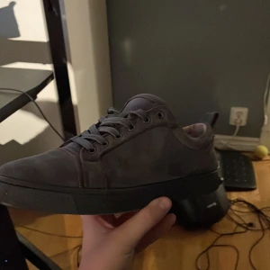 Grå sneakers med röd sula - Snygga grå sneakers från Christian Louboutin med den ikoniska röda sulan. Skorna har snörning och är tillverkade i suede, vilket ger en lyxig känsla. Perfekta för dig som vill ha en stilren look med en touch av exklusivitet. Passar 41-42