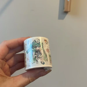Dekorativ tejp med miku hatsune  - Säljer en rulle dekorativ tejp med ett färgglatt anime-motiv. Tejpen har en vit bas med illustrationer av en karaktär med långt blått hår och olika små detaljer som ger en lekfull känsla. Perfekt för att dekorera anteckningsböcker eller paket.