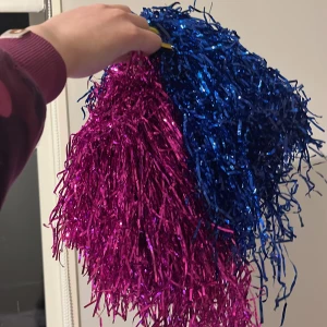 Blå och rosa pom poms - Snygga pom poms i glittrande blått och rosa, perfekta för att heja på ditt lag eller för en festlig dansuppvisning. De har ett gult handtag för ett bekvämt grepp.