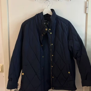Mörkblå quiltad jacka från Lindex  - Snygg mörkblå quiltad jacka från Lindex med guldknappar och hög krage. Jackan har långa ärmar och praktiska fickor framtill. Perfekt för kyligare dagar.