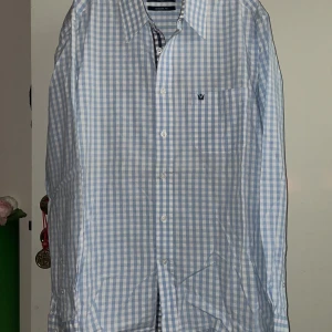 Melka of Sweden rutig skjorta  - Snygg blå och vit rutig skjorta från Melka of Sweden i modern passform. Skjortan har långa ärmar och knappar framtill. Perfekt för en stilren look med ett diskret broderat emblem på bröstfickan.