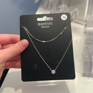 Silverfärgat halsband från Montini - Elegant silverfärgat halsband från Montini med en enkel kedja och en gnistrande sten som hänge. Perfekt för att lägga till en touch av glamour till din outfit.