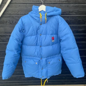Blå dunjacka från Fjällräven - Säljer en blå dunjacka från Fjällräven med huva och gula detaljer. Jackan har knappar och dragkedja framtill samt praktiska fickor med knappar. Perfekt för kyliga dagar. Den har ett pyttelitet hål som lagats, det sitter under ena ärmen och syns på fjärde bilden. Det är så litet så man kan behöva zooma in för att se det.