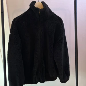 Svart teddyjacka - Mysig svart teddyjacka med dragkedja och hög krage. Perfekt för kyliga dagar när du vill hålla dig varm och bekväm. Jackan har en avslappnad passform och långa ärmar.