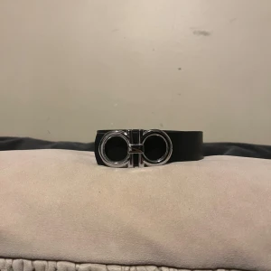 Svart ferragamo bälte - Snyggt svart bälte med en stilren silverfärgad spänne i metall. Perfekt för att ge en elegant touch till din outfit. Passar både till jeans och kostymbyxor.