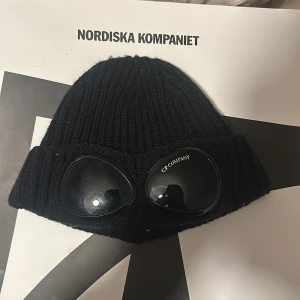 Svart stickad mössa från C.P. Company - Snygg svart stickad mössa från C.P. Company med inbyggda glasögon. Perfekt för att hålla stilen under kalla dagar. Mössan har en ribbad design och är både trendig och funktionell.