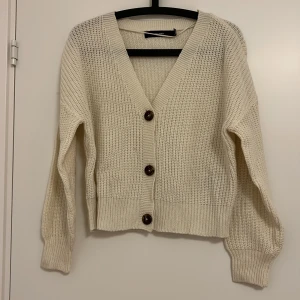 Vit stickad kofta från Vero Moda - Säljer en stilren vit stickad kofta från Vero Moda. Koftan har en v-ringad design med stora knappar framtill och långa ärmar. Perfekt för lager-på-lager under kyligare dagar.