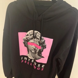 Svart hoodie med tryck - Cool svart hoodie med ett konstnärligt tryck av en staty och texten 'Intense Feelings' i rosa och vitt. Hoodien har en justerbar dragsko i huvan och en bekväm passform. Kan vara lite nopprig 