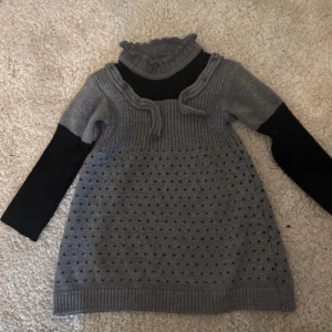 Grå stickad klänning med svarta ärmar - Säljer en grå stickad klänning med svarta långärmade inslag. Klänningen har en ribbad polokrage och ett dekorativt hålmönster på framsidan. Perfekt för kyligare dagar.