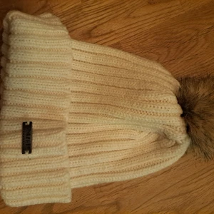 Vit stickad mössa med pälsboll - Snygg vit stickad mössa från j lindberg med en fluffig pälsboll på toppen. Perfekt för att hålla värmen under kalla dagar. Mössan har en ribbad design och en metallplatta med märkeslogga framtill.