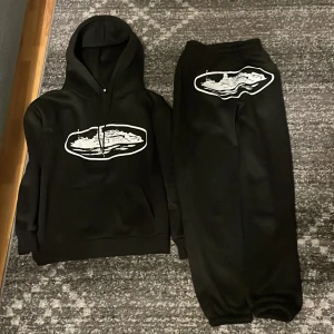 Svart cortize tracksuit - Säljer en svart cortize tracksuit. Perfekt för en avslappnad stil och passar bra till både vardag och chill. Hoodien har en klassisk passform med långa ärmar och en skön huva.