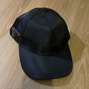 Svart keps från Prada - Snygg svart keps från Prada i bomull. Den har en stilren design med en justerbar rem baktill för perfekt passform. Perfekt för både vardag och fest! 🖤