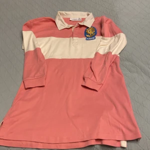 Rosa och vit randig klänning från Mini Rodini - Säljer en superfin rosa och vit randig pikénlänning från Mini Rodini. Den har en cool blomma med texten 'M. Rodini' på bröstet. Klänningen är i mycket bra skick, använd vid ett tillfälle, och är gjord av ekologisk bomull. Passar perfekt för en avslappnad stil och passar perfekt med stickade strumpbyxor! 