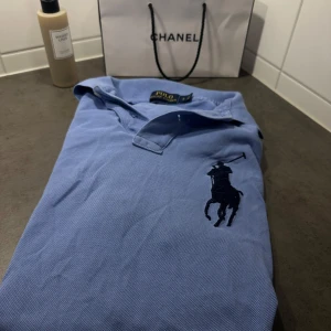 Blå pikétröja från Polo Ralph Lauren - Säljer en snygg blå pikétröja från Polo Ralph Lauren i storlek S. Tröjan har en klassisk krage och korta ärmar med en stor broderad logga på bröstet. Perfekt för en avslappnad men stilren look. Passar både till vardags och fest!