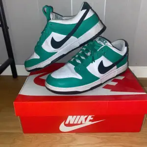 Säljer ett par Nike Dunk sneakers i grönt och vitt. De har en klassisk design med svart swoosh och snörning. Perfekta för både vardag och träning. Skorna är i bra skick och kommer i originalkartong. De är helt rena ocj fräscha kommer med 2 par crease skydd